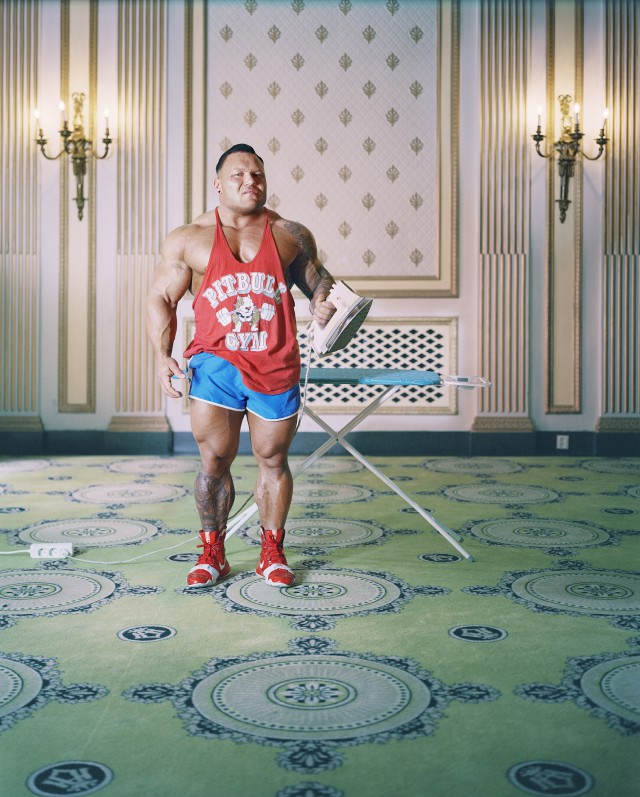Bodybuilder stands with an iron in a hotel room. The bodybuilder is wearing shorts and a tank top with a logo.