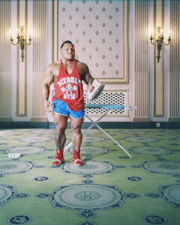 Bodybuilder stands with an iron in a hotel room. The bodybuilder is wearing shorts and a tank top with a logo.
