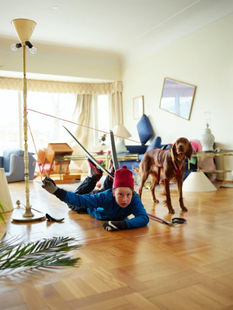 A child in a red hat and blue jacket lies on the floor with a dog and makeshift ski equipment in a brightly lit living room.