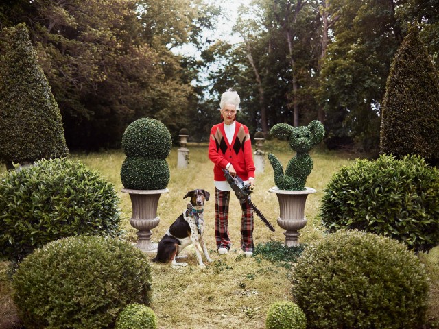 Adult woman in a red cardigan using a chainsaw near a Dalmatian mix dog in a lush outdoor garden surrounded by topiary bushes.