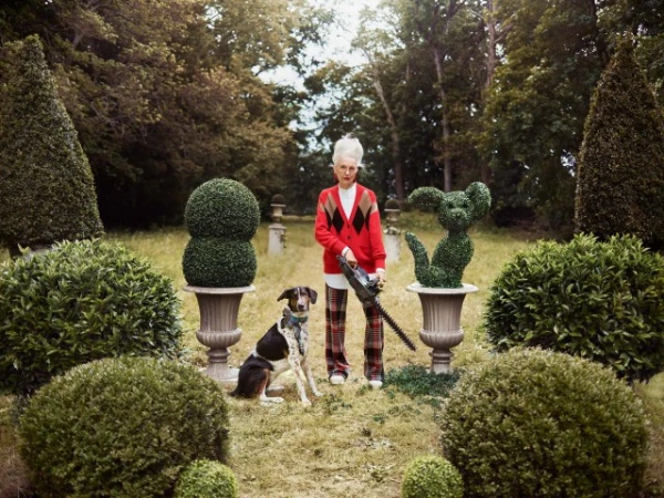 Adult woman in a red cardigan using a chainsaw near a Dalmatian mix dog in a lush outdoor garden surrounded by topiary bushes.