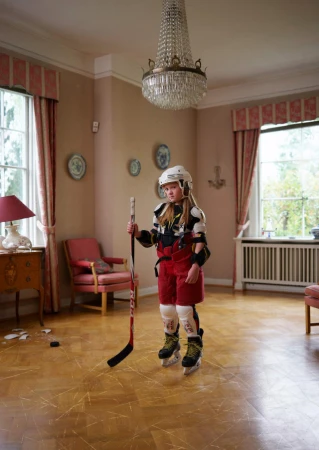 Young girl in hockey gear stands indoors with stick near broken decor. Chandelier hangs from ceiling, natural light streams through windows.
