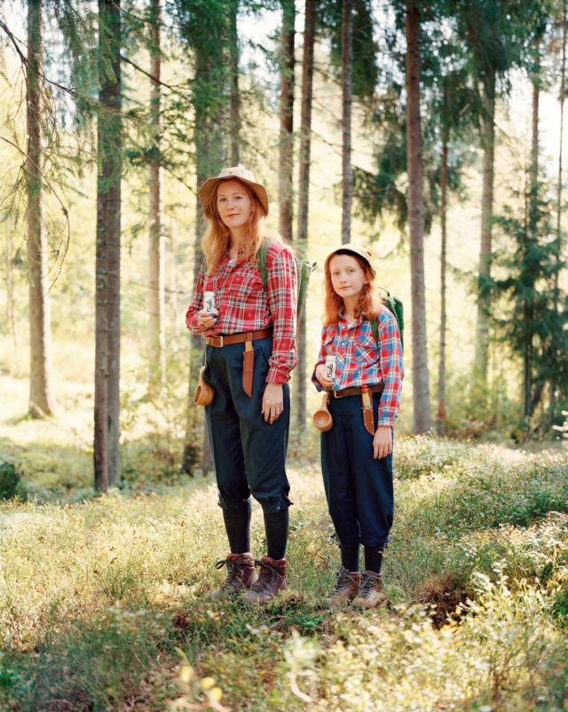 Two people, an adult and a child, wearing plaid shirts and hats hiking in a forest with tall trees and green ground cover.