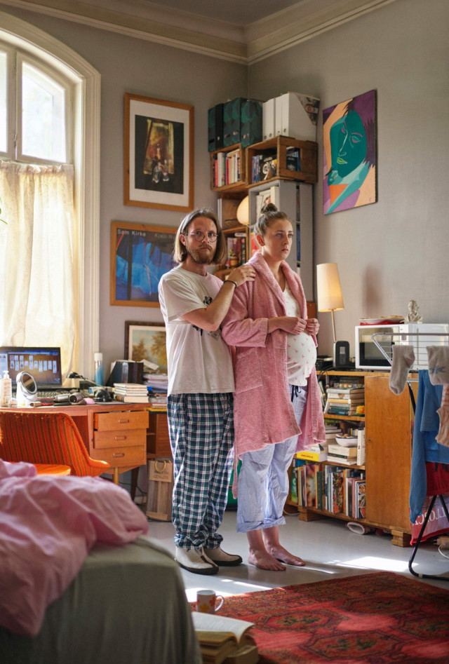 A pregnant woman stands indoors with a man; she's in a pink robe, while he wears pajamas; a bookshelf and orange desk fill the room.