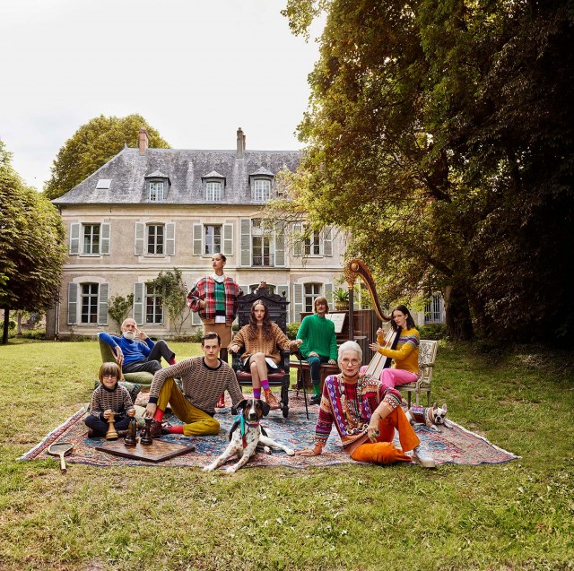 Group of people of various ages gather on a rug in a grassy yard near a stately home, with a dog, chess set, and musical instruments.