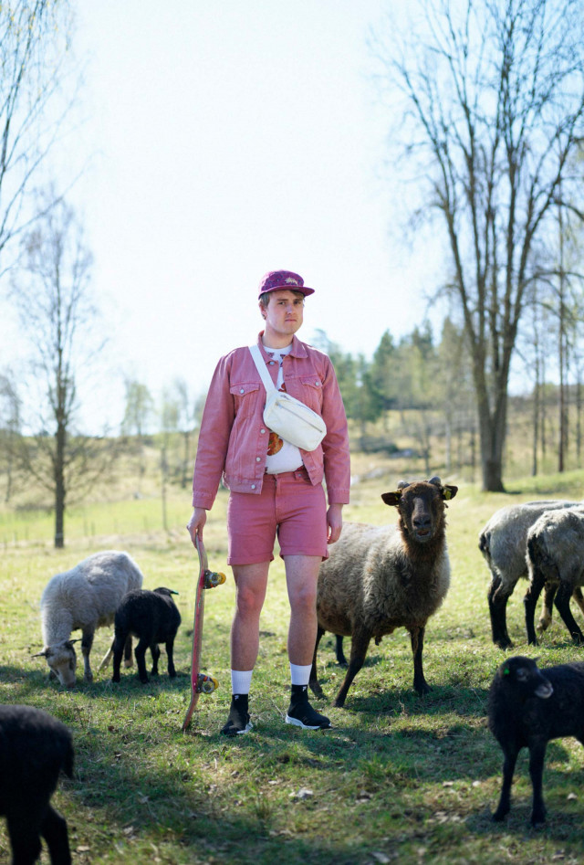 An adult male stands in a field of sheep, wearing a pink outfit with a skateboard and a fanny pack on a bright day.
