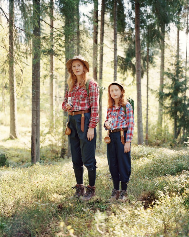 Woman and girl hiking in a forest, wearing hats and plaid shirts, with trees and sunlight creating a natural, outdoor setting.