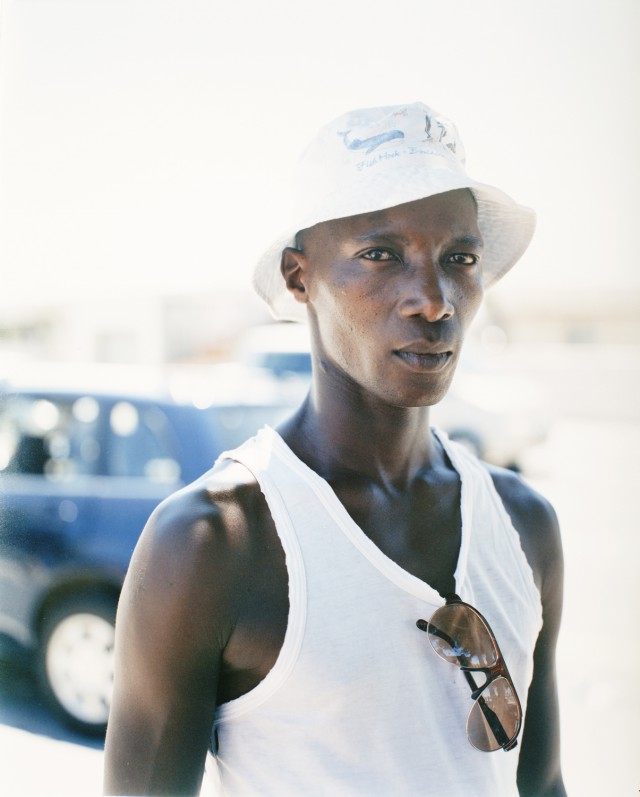 Black man wearing a white tank top and a white bucket hat with sunglasses tucked into the neckline