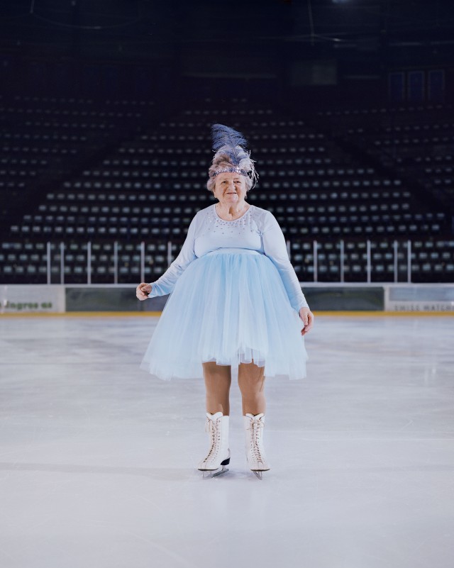 Elderly woman ice skating on an ice rink. She is wearing a blue dress, ice skates and a blue feather headpiece.