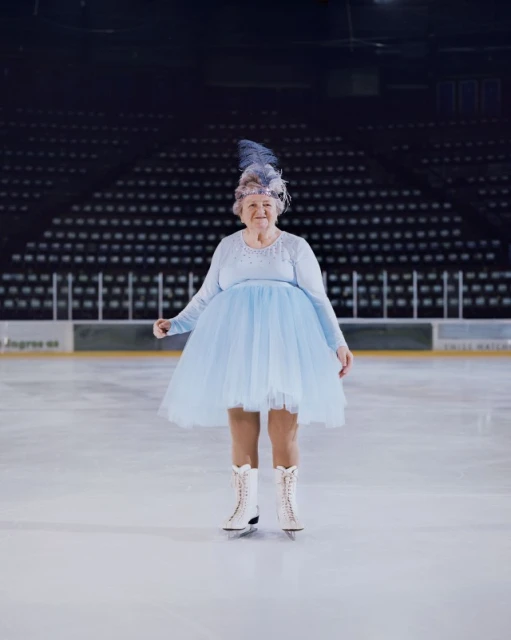 Elderly woman ice skating on an ice rink. She is wearing a blue dress, ice skates and a blue feather headpiece.