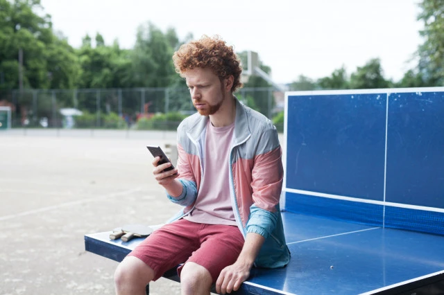 Man with red hair sits on a blue ping pong table outdoors using his smartphone, wearing a colorful jacket and red shorts on a sunny day.