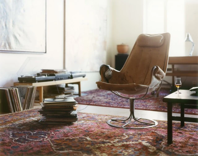 A vintage leather chair sits on a patterned rug in a living room with a record player and vinyl records. Natural light illuminates the room.