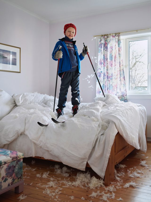 Boy smiles standing on a bed covered in feathers wearing skis and holding ski poles in a brightly lit bedroom with floral curtains.