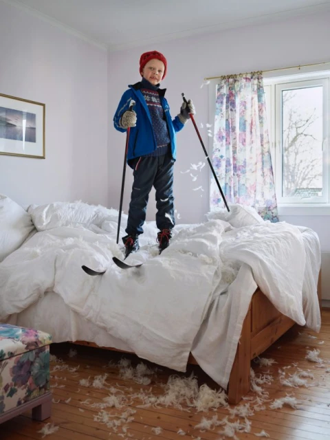 Boy smiles standing on a bed covered in feathers wearing skis and holding ski poles in a brightly lit bedroom with floral curtains.