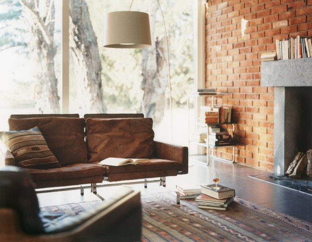 Cozy living room with brown sofa, books, and brick fireplace. Natural light streams through large windows, enhancing the warm interior.