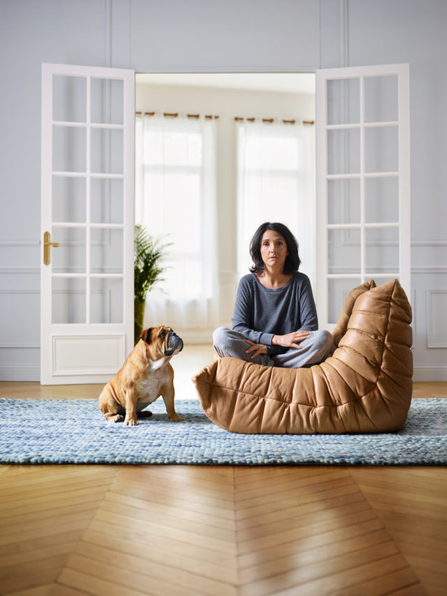 Woman in grey loungewear sits cross-legged on tan leather beanbag chair indoors with a bulldog on a blue rug near open french doors.