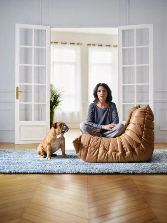 Woman in grey loungewear sits cross-legged on tan leather beanbag chair indoors with a bulldog on a blue rug near open french doors.