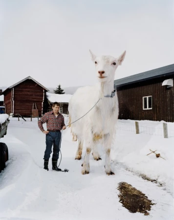 Full shot of a man standing with a white goat on a snowy path near rustic wooden buildings in a rural winter setting under an overcast sky.