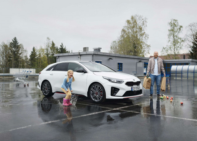 A young girl in pink boots jumps in a puddle with a white car and an adult male holding shopping bags nearby in an outdoor parking lot.