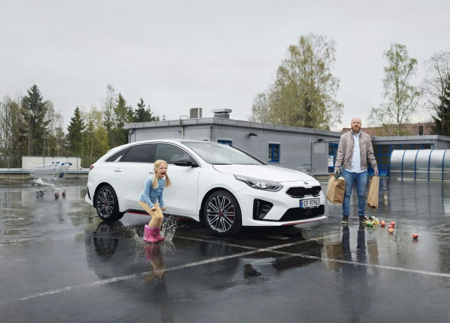 A young girl in pink boots jumps in a puddle with a white car and an adult male holding shopping bags nearby in an outdoor parking lot.