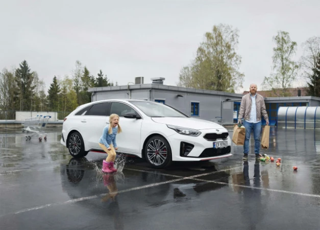A young girl in pink boots jumps in a puddle with a white car and an adult male holding shopping bags nearby in an outdoor parking lot.