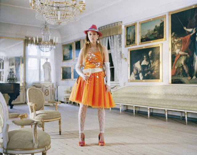 A woman in an orange dress, pink hat, and matching heels stands in a formal room with paintings and chandeliers, holding a glass.