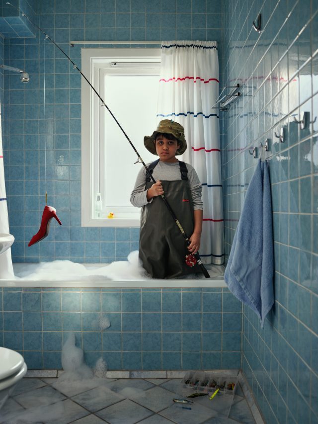Child in overalls and a bucket hat fishing from a bubble bath with a red high heel hanging from the fishing line in a blue tile bathroom.