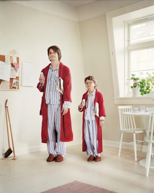 Two individuals, one adult male and one child, stand indoors wearing matching striped pajamas and red robes, holding books.