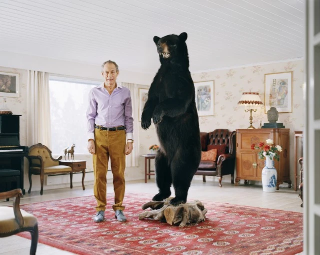 Eye-level full shot of a man standing next to a taxidermied bear in a light-filled room with a patterned rug and wallpaper.