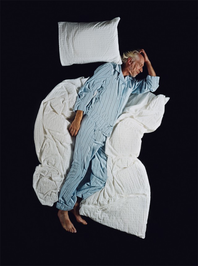 Overhead shot of an adult male sleeping on a white wrinkled blanket with a pillow on a black background in blue and white striped pajamas.