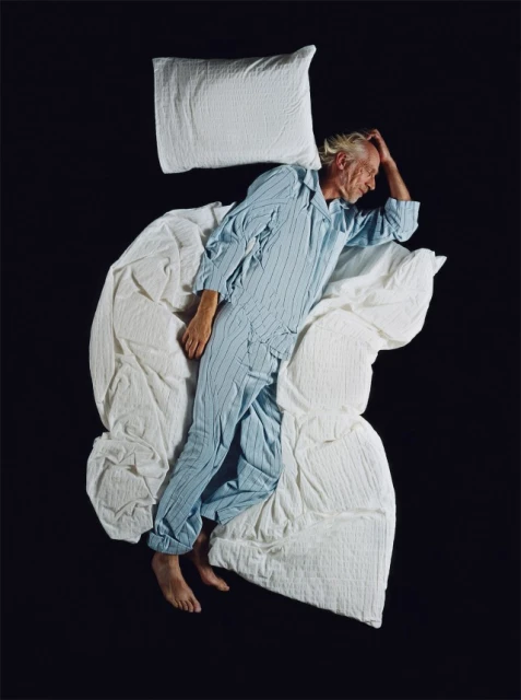 Overhead shot of an adult male sleeping on a white wrinkled blanket with a pillow on a black background in blue and white striped pajamas.