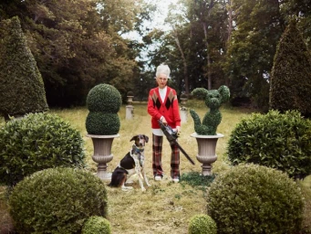 Elderly woman holding a hedge trimmer in a formal garden with a dog and topiary