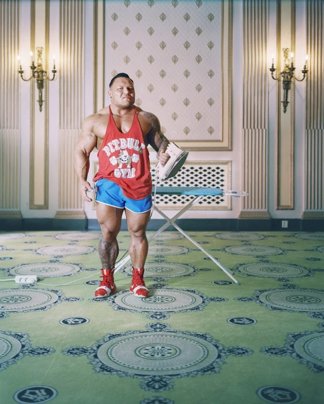 Muscular man wearing a red tank top and blue shorts holding an iron standing in an ornate room photographed by Morten Borgestad