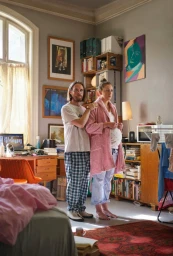 Man standing behind pregnant woman wearing a pink robe inside a cluttered domestic living room