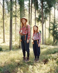 Woman and child wearing matching plaid shirts and hats standing in a sunlit pine forest.