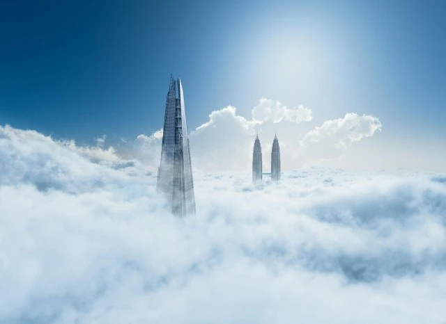 Skyscrapers pierce through a sea of clouds beneath a blue sky. Two distant towers and a taller, modern building are visible.