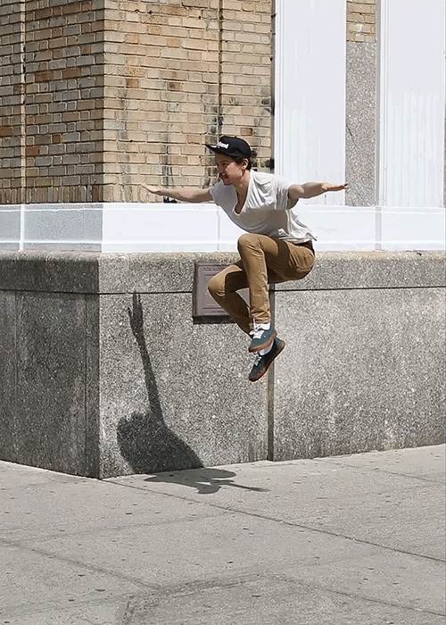 Active man in brown pants and white t-shirt mid-air jump with arms outstretched, capturing urban movement and energy against city backdrop.