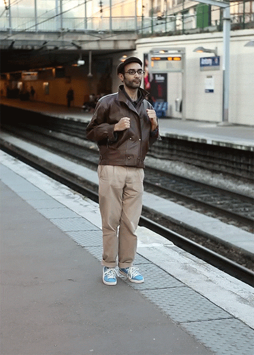 Man in brown leather jacket standing on train platform in an urban setting with railway tracks and station signage.