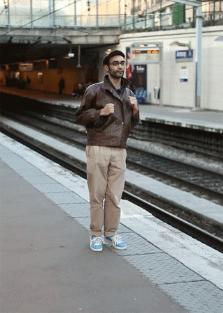 Man in brown leather jacket standing on train platform in an urban setting with railway tracks and station signage.