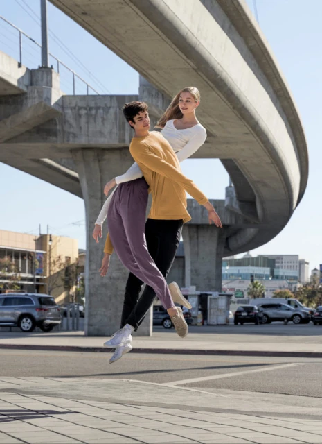 Two dancers leap outdoors under a concrete bridge, wearing long-sleeved tops and pants, capturing a blend of urban and artistic expression.
