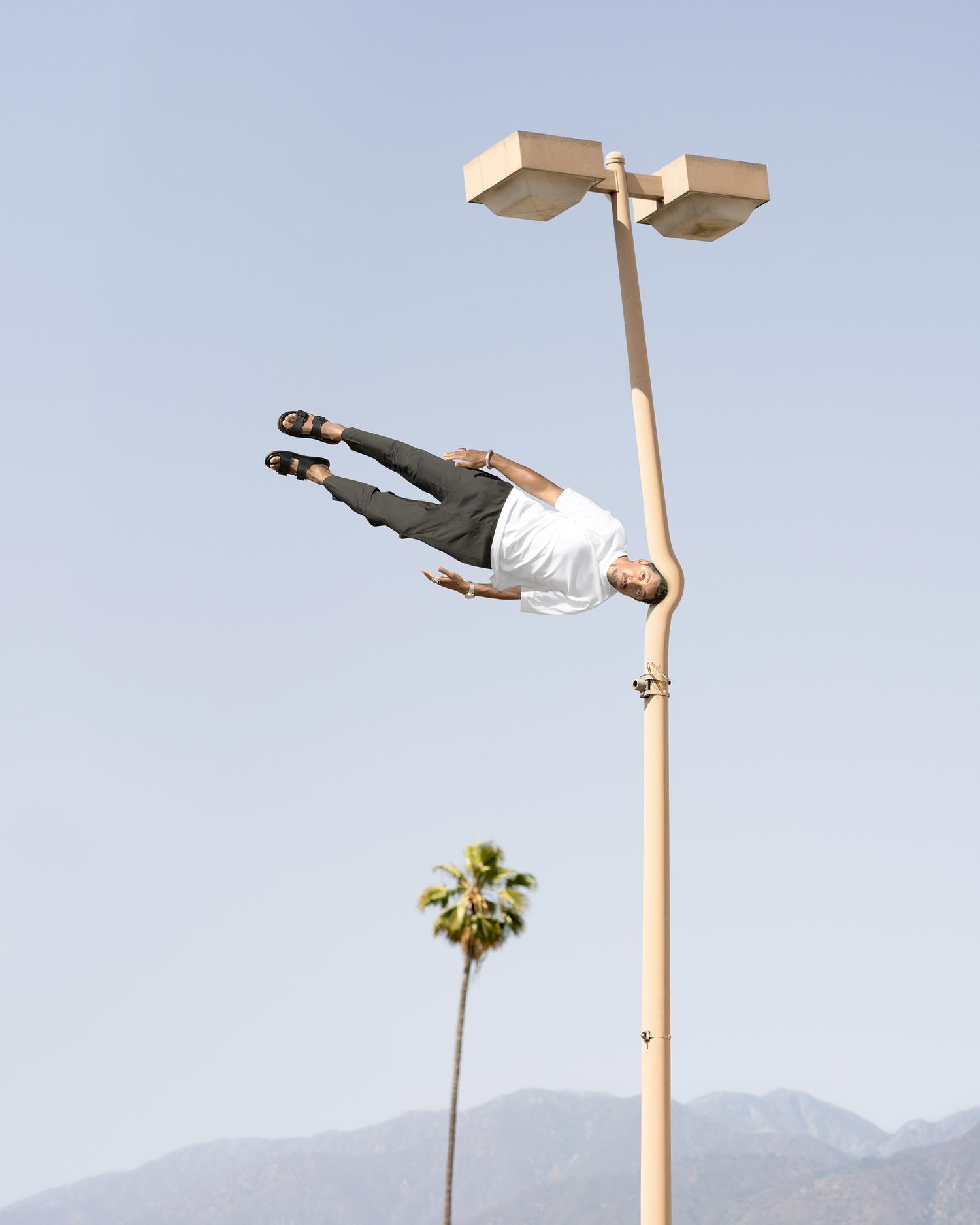 An adult male is doing a handstand on top of a beige streetlight on a sunny day with a palm tree and mountains in the background.
