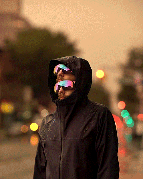 Adult man in a hooded black raincoat wears futuristic, colorful sunglasses in an urban street setting with blurred city lights.