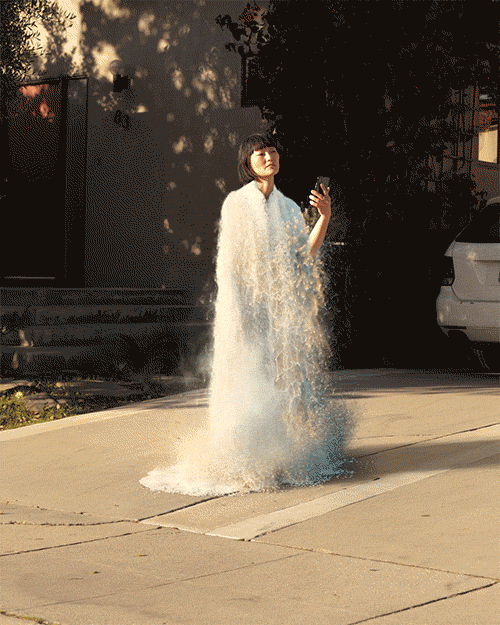 Woman standing on sidewalk in digitally rendered dress, holding cellphone, bathed in sunlight, urban street, suburban home in background.