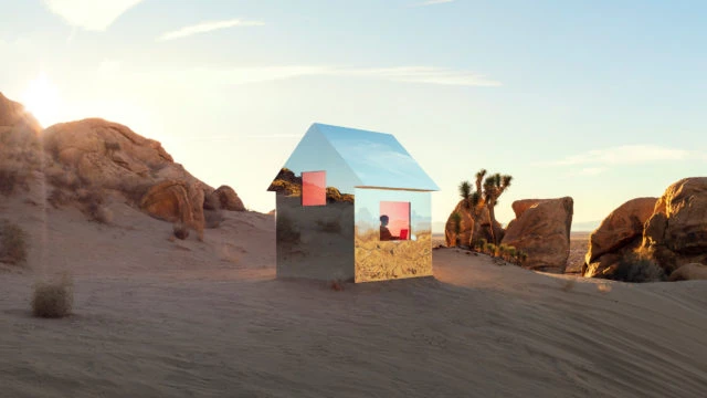 Mirrored house in a desert landscape, reflecting the surrounding environment and an individual visible through a window, under a blue sky.