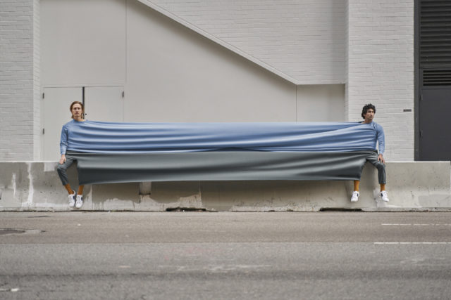 Two adults sit on a concrete barrier holding a large blue and gray fabric outdoors, on a city street with a concrete wall backdrop.