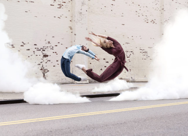 Two adults perform a synchronized dance, arching back against a brick wall with smoke effects and urban asphalt ground in artistic photo.
