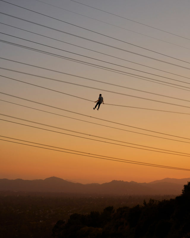 A person suspended from power lines against an orange and blue gradient sky, silhouetted hills visible in the distance.