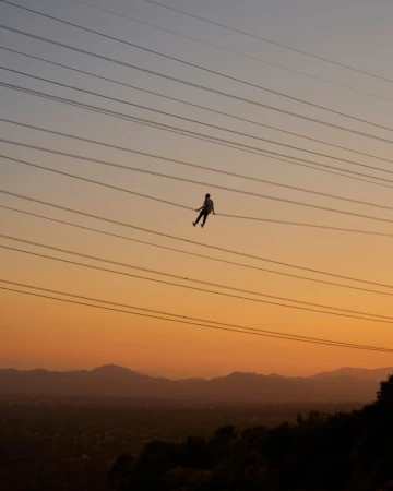 A person suspended from power lines against an orange and blue gradient sky, silhouetted hills visible in the distance.