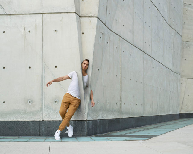 Adult male dancer in a white tee and tan pants leans against an angled concrete wall outdoors in an urban setting, captured in medium shot.