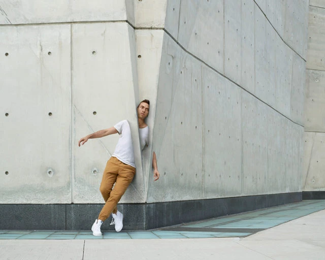 Adult male dancer in a white tee and tan pants leans against an angled concrete wall outdoors in an urban setting, captured in medium shot.